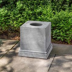 a concrete trash can sitting on top of a sidewalk next to some bushes and trees