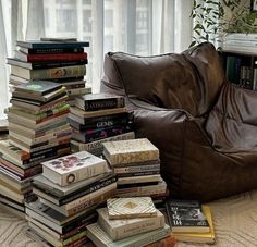 a pile of books sitting on the floor next to a brown leather chair in front of a window