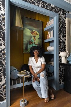 a woman sitting on the floor in front of a blue book shelf with a painting behind her