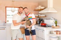 two men standing in a kitchen holding a cat