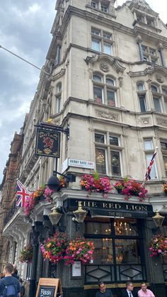 people are walking in front of a building with flowers on the outside and an awning above it