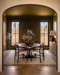 a dining room table with four chairs and a vase on top of it in front of two windows