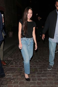 a woman in black shirt and jeans walking on brick road with people behind her at night