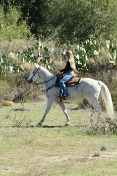 a woman riding on the back of a white horse in a field next to trees