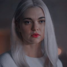 a woman with long white hair and red lipstick looks into the camera while wearing a white sweater