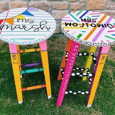 two colorful stools sitting in the grass near a brick wall with writing on them