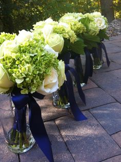 three vases with flowers and blue ribbon on the side of a brick walkway in front of trees