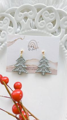 small christmas tree earrings on a white plate with red berries in the foreground and an ornamental design behind it