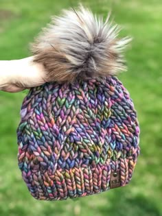 a hand holding a multicolored knitted beanie with a fuzzy pom - pom