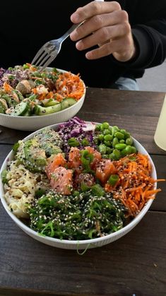 two bowls filled with different types of food on top of a wooden table next to a person holding a fork