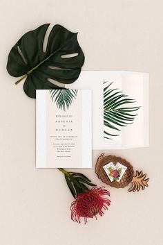 the wedding stationery is laid out with tropical leaves and greenery, including a red flower