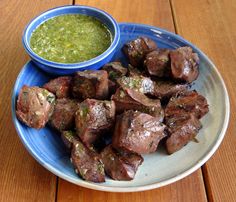 a blue plate topped with meat next to a bowl of green sauce on top of a wooden table