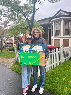 two people standing next to each other in front of a house holding a sign that says,