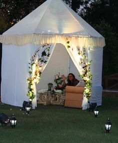 a woman sitting in a chair under a white tent with flowers and greenery on it