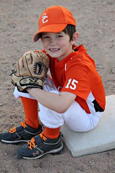 a young boy in an orange baseball uniform is sitting on the ground with his glove