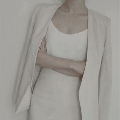 a woman wearing a white dress and jacket with her arms folded over her chest, standing in front of a wall