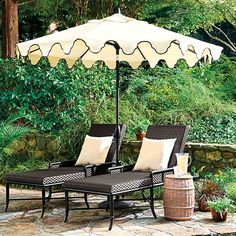 two lounge chairs under an umbrella on a stone patio with plants and trees in the background