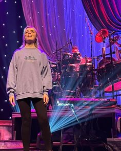 a woman standing on top of a stage next to a drum set with lights behind her