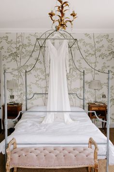 a white dress hanging on a bed in a room with floral wallpaper and chandelier