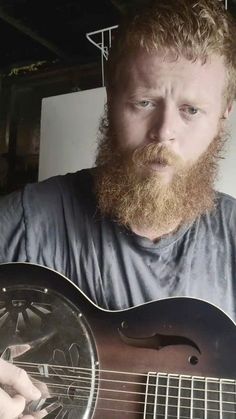 a man with a beard is holding a guitar and looking at the camera while wearing a blue shirt