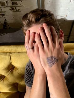 a man covering his face with both hands while sitting on a yellow couch in front of a brick wall