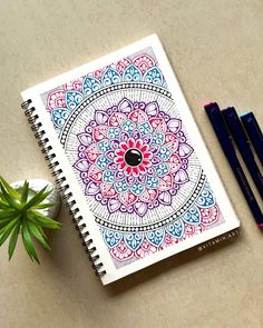 a notebook with an intricate design next to two markers and a potted plant on a table