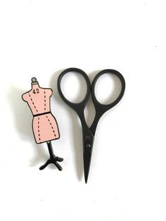 a pair of scissors and a pin with a pink dress on it sitting next to each other