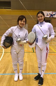 two women in fencing uniforms holding their racquets on a wooden floor with an indoor court