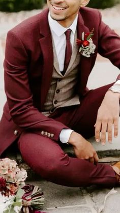 a man in a red suit and tie sitting on the ground with flowers around him