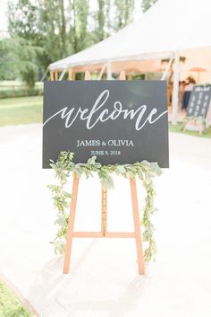 a welcome sign with greenery on it in front of a tent at a wedding