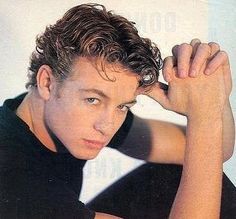 a young man with curly hair and black shirt posing for a photo in front of a white background