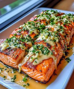 salmon covered in cheese and herbs on a white platter next to a glass window