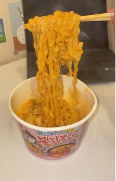 noodles being lifted from a bowl with chopsticks in front of a laptop computer