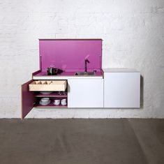 a white and pink kitchen with drawers on the side, sink and countertop in front of a brick wall