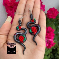 a pair of black and red snake earrings with roses on the ends, in front of pink flowers