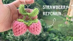 a hand holding two crocheted strawberry keychains in front of a forest