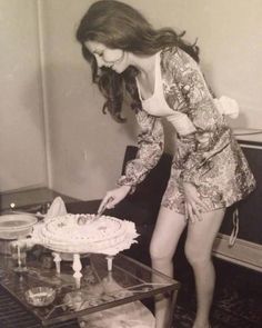 a woman cutting a cake on top of a glass table in front of a fireplace