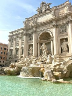 a fountain in front of a large building with statues on it's sides and water running down the side