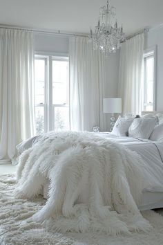 a white bedroom with a chandelier hanging from the ceiling and fluffy blankets on the bed