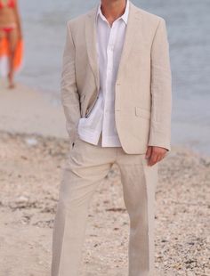 a man standing on top of a sandy beach next to the ocean wearing a white shirt and tan suit