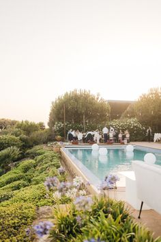 an outdoor pool with people sitting at it and flowers in the foreground, surrounded by greenery