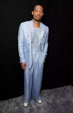 a man standing in front of a black backdrop wearing a blue suit and white shoes