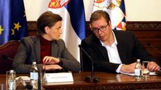 two people sitting at a table in front of flags and microphones, with one person holding a pen