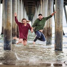 two people jumping into the water under a pier with their arms in the air and smiling