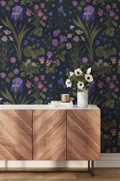 a vase with flowers on top of a wooden cabinet in front of a floral wallpaper