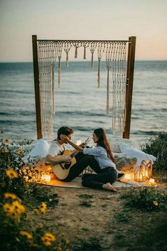 a man and woman sitting on the ground playing guitar next to an outdoor fire pit