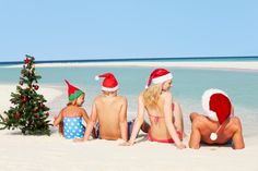 three people in santa hats sitting on the beach next to a christmas tree and water