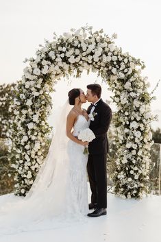 a bride and groom kissing in front of an arch of flowers at their winter wedding