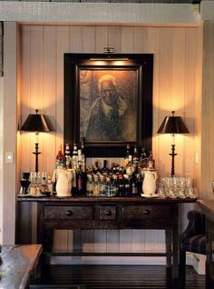 a room with two lamps and a painting on the wall next to a table full of liquor bottles