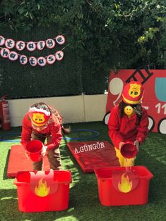 two children in fireman costumes playing with buckets on fake grass at an outdoor birthday party